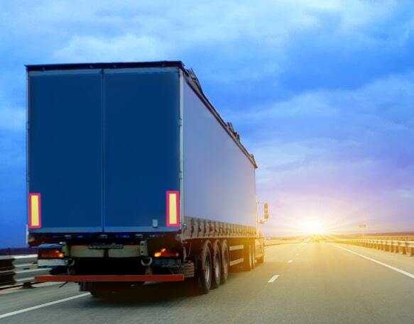 A blue semi-truck drives down the highway toward a shining sunset.