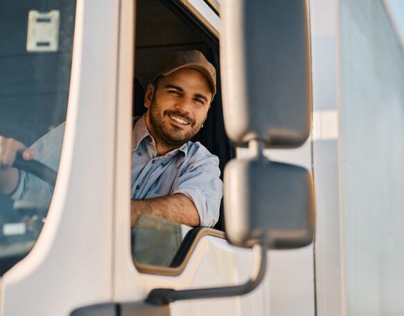 A smiling truck driver holds the steering wheel while sitting in the driver's seat of a semi truck and leaning slightly out the window.