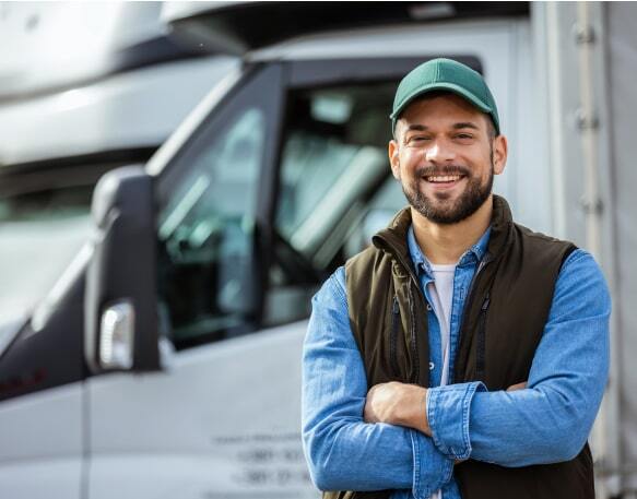 Smiling truck driver in a denim shirt and brown vest standing in front of a semi truck.