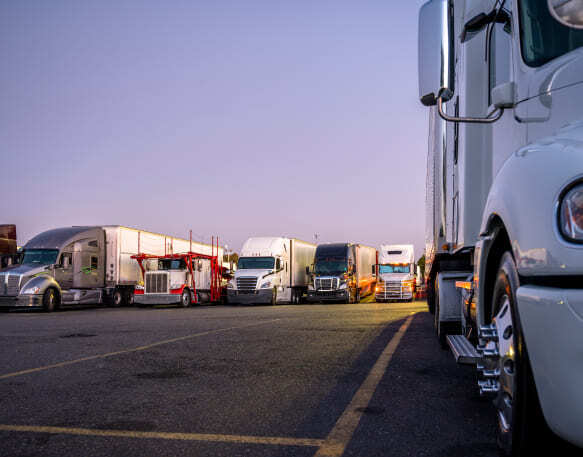 A line of semi trucks with trailers parked in a spacious parking lot at dusk, with one truck parked closer in the foreground.