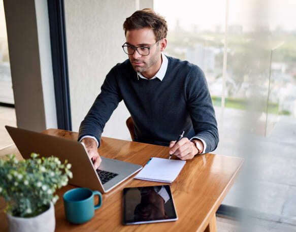 Young businessman sitting at a table at home working on a laptop and writing down ideas in a notebook.