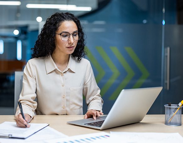 A female shipper works hard while looking at laptop and writing on a piece of paper inside her office.