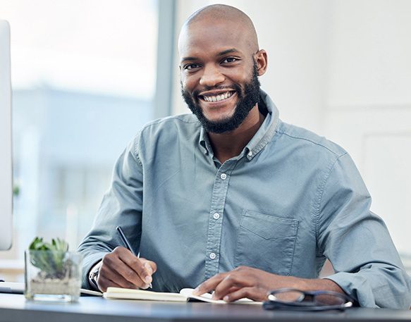 Male shipper in the trucking industry working at his desk and writing in a notepad while smiling at the camera.