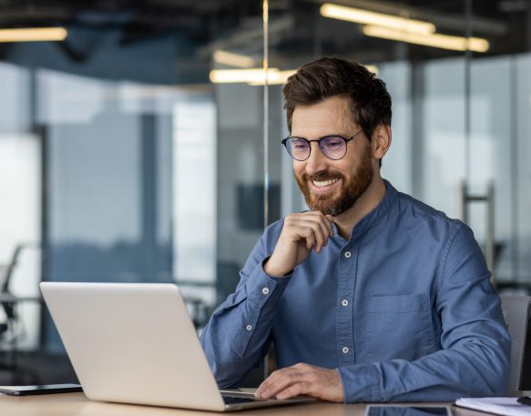 A young man in glasses and a denim shirt is sitting in the office at a desk, working in the shipper industry and talking on a video call online.