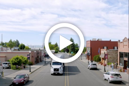 Play button overlay on an image showing a semi-truck driving down a small town street, with buildings and parked cars lining both sides.